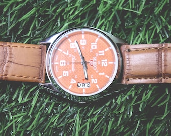 Relógio masculino Seiko 5 automático vintage, mostrador laranja, caixa prateada, pulseira de couro marrom.