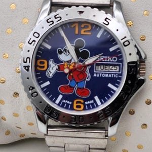 Puede incluir: Un reloj de pulsera automático Seiko plateado con una esfera azul con un gráfico de Mickey Mouse. El reloj tiene una correa de acero inoxidable y una pantalla de fecha. La esfera del reloj tiene números y agujas naranjas.