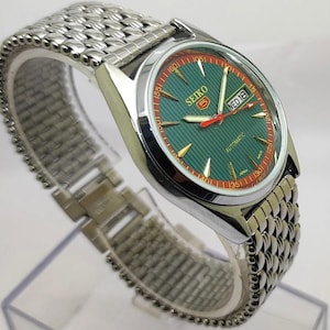 May include: A silver-toned Seiko 5 automatic wristwatch with a green dial and a red and gold bezel. The watch has a silver mesh band and a day-date display. The watch face displays the Seiko logo and the word "Automatic".
