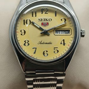 以下が含まれることがあります： イエローの文字盤が付いた、シルバーのセイコー自動巻き腕時計。文字盤には黒い数字、針、曜日が表示されています。ステンレススチールのバンドが付いています。