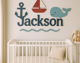 May include: A wall-mounted wooden name sign reading Jackson in dark blue letters, accompanied by a dark blue anchor, a red and white sailboat, a light blue whale, and a wavy light blue line, all arranged above a white wooden crib.