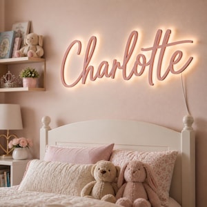 May include: A pink, illuminated 'Charlotte' name sign in a cursive font is mounted on a pink wall above a white bed. The sign has a warm glow. The bed is made with pink and white bedding, and there are stuffed toys on the bed.