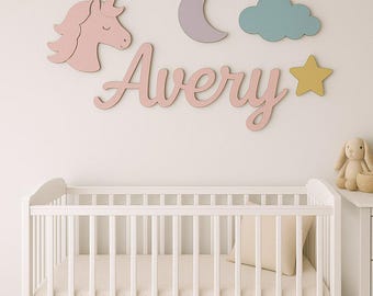 May include: A wall-mounted wooden name sign displaying the name Avery in pink cursive script, accompanied by a pink unicorn head, a lavender crescent moon, a teal cloud, and a yellow star, all arranged as decorative wall cutouts above a white crib.