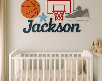 Könnte beinhalten: Ein Wanddekorationsset bestehend aus einem Basketball, einem Basketballkorb, einem hellblauen Stern, einem grauen Sneaker und dem Namen Jackson in dunkelblauer Schreibschrift, angebracht über einem weißen Kinderbett.