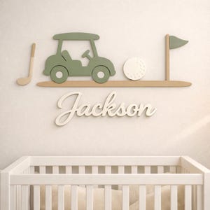 May include: A wall-mounted wooden sign featuring a sage green golf cart, a white golf ball, a tan golf club, and a flag on a tan base. Below the golf elements, the name Jackson is written in white cursive script lettering on the wall.