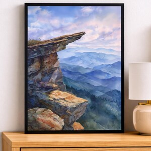 May include: A framed wall art print featuring a watercolor painting of a rocky cliff edge overlooking layers of blue mountain ranges under a soft, pastel-colored sky with light clouds, displayed in a simple black frame on a wooden surface.