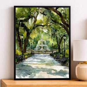 Op de afbeelding: Aquarelprint van een parkscène met een fontein, banken en bomen. Het kunstwerk is zwart omlijst en wordt getoond op een houten nachtkastje. Een lamp met een witte kap staat in de buurt.