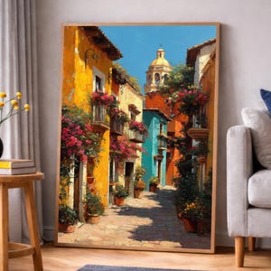 May include: A framed art print depicting a vibrant street scene. Buildings in shades of yellow, orange, and turquoise line the cobblestone street, adorned with balconies overflowing with pink flowers. A domed structure is visible in the background under a clear blue sky.