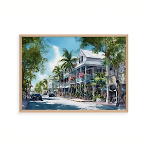 May include: Watercolour painting of a tropical street scene with buildings, palm trees, and cars. The artwork is framed in a light wood frame. The buildings have balconies with pink flowers. The sky is blue with white clouds.