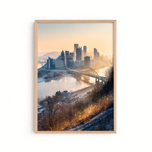 May include: A framed print of a cityscape featuring a river, bridges, and skyscrapers. The image has a warm color palette with a golden sky and a light brown wooden frame. The city skyline is silhouetted against the sky.