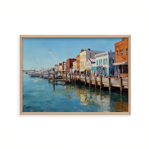 May include: An oil painting in a light wood frame depicting a harbour scene with a wooden pier extending into blue water. A row of multi-storey buildings in shades of orange, yellow, and white lines the dock, with several sailboats moored nearby.