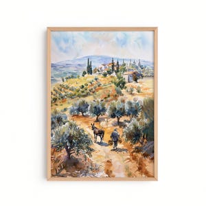 May include: Watercolor painting of a rural landscape with olive trees, a donkey, and a person walking on a path. The artwork is framed in a light wood frame. The color palette includes greens, browns, and blues, depicting a sunny day.