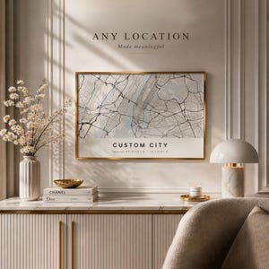 Op de afbeelding: Een ingelijste kaartafdruk met een gouden rand, getiteld "CUSTOM CITY", hangt boven een witte kast. De tekst "ANY LOCATION Made meaningful" staat boven de kaart. Een witte vaas met bloemen, boeken en een lamp staan op de kast.