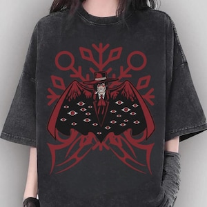May include: Dark gray t-shirt featuring a red graphic of a character with a hat, coat, and multiple eyes. The design includes a snowflake-like pattern and decorative elements. The shirt has short sleeves and a relaxed fit.