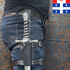May include: A silver-toned dagger with a spiralled black and silver handle secured in a black leather thigh holster. The holster features adjustable straps with metal buckles and rivets, worn over dark blue denim jeans with a black leather belt.
