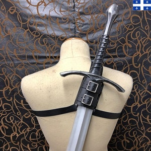 Puede incluir: Una espada de estilo medieval con una hoja plateada y un mango envuelto en cuero negro. La espada está sujeta a un maniquí con un arnés de cuero negro. El fondo es un patrón oscuro.