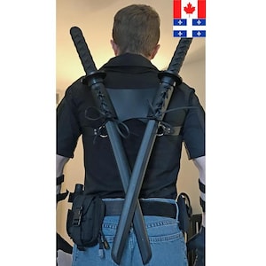 Op de afbeelding: Een persoon draagt een zwart shirt en een blauwe jeans met een zwart leren harnas en twee zwarte zwaarden die op hun rug zijn vastgemaakt.