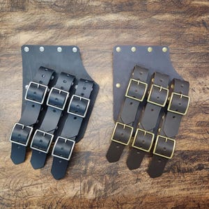 Peut inclure: Deux lanières en cuir avec plusieurs boucles. Les lanières sont brunes et noires et ont des boucles argentées et dorées.