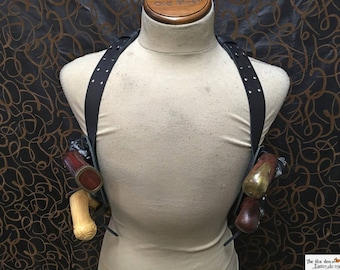 May include: A black leather holster with two silver-toned pistols. The holster has a strap that goes over the shoulder and is decorated with silver rivets.