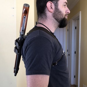 May include: A person wearing a black t-shirt and a leather shoulder strap with a rifle attached to it. The rifle has a wooden stock and a metal barrel. The strap is secured with a buckle. The person is standing in a hallway with a door in the background. The text "The fox den L'antre du renard" is visible in the bottom right corner of the image.