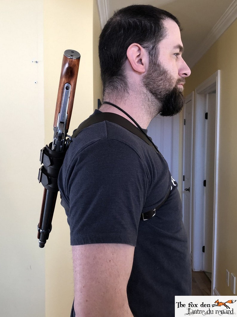 May include: A person wearing a black t-shirt and a leather shoulder strap with a rifle attached to it. The rifle has a wooden stock and a metal barrel. The strap is secured with a buckle. The person is standing in a hallway with a door in the background. The text "The fox den L'antre du renard" is visible in the bottom right corner of the image.