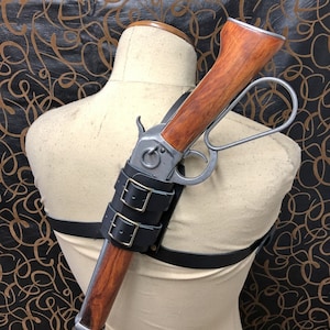 May include: A leather shoulder holster with a rifle attached. The holster has two buckles and a strap that goes over the shoulder. The rifle has a wooden stock and a metal barrel.
