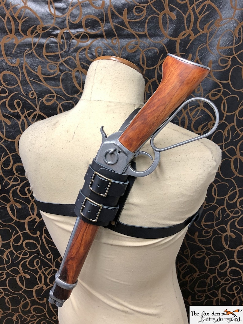May include: A leather shoulder holster with a rifle attached. The holster has two buckles and a strap that goes over the shoulder. The rifle has a wooden stock and a metal barrel.