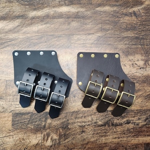 Peut inclure: Deux boucles de ceinture en cuir pour épée avec trois boucles chacune. La boucle de gauche a des boucles argentées, et la boucle de droite a des boucles dorées.