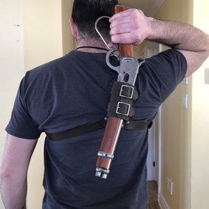 May include: A person wearing a dark grey t-shirt is wearing a leather shoulder holster with a silver and brown rifle attached. The rifle is secured with two leather straps.