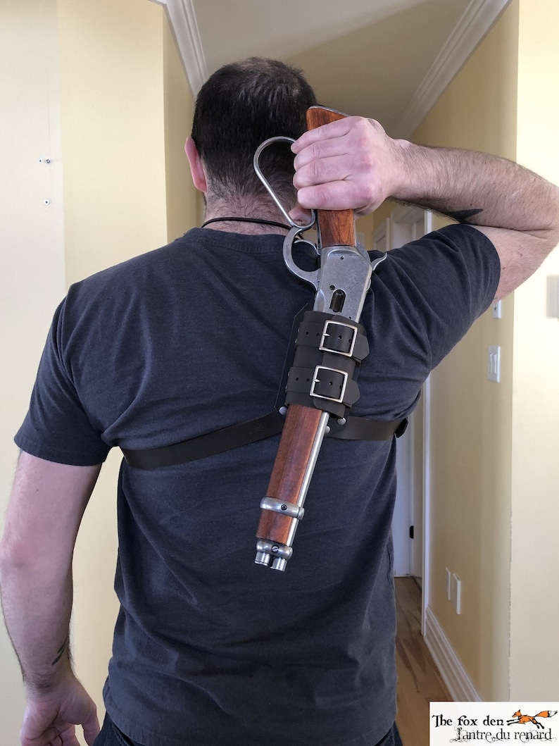 May include: A person wearing a dark grey t-shirt is wearing a leather shoulder holster with a silver and brown rifle attached. The rifle is secured with two leather straps.