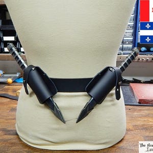 May include: A black leather belt with two black leather sheaths for holding daggers. The daggers have black handles and silver blades. The belt is worn on a white mannequin.