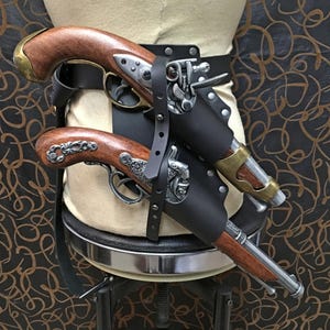 Peut inclure: Deux pistolets de style antique avec des poignées en bois et des accents métalliques sont fixés dans un holster en cuir noir. Le holster est attaché à une ceinture en cuir marron.