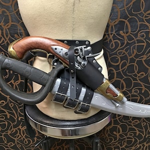 Peut inclure: Une ceinture en cuir noir avec une boucle argentée et deux étuis. Un étui contient un pistolet plaqué argent avec une poignée en bois, et l'autre étui contient une dague plaquée argent avec une poignée noire.