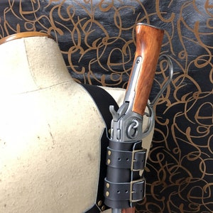 May include: A black leather gun holster with two buckles and brass studs. The holster is attached to a mannequin and holds a long gun with a wooden stock.