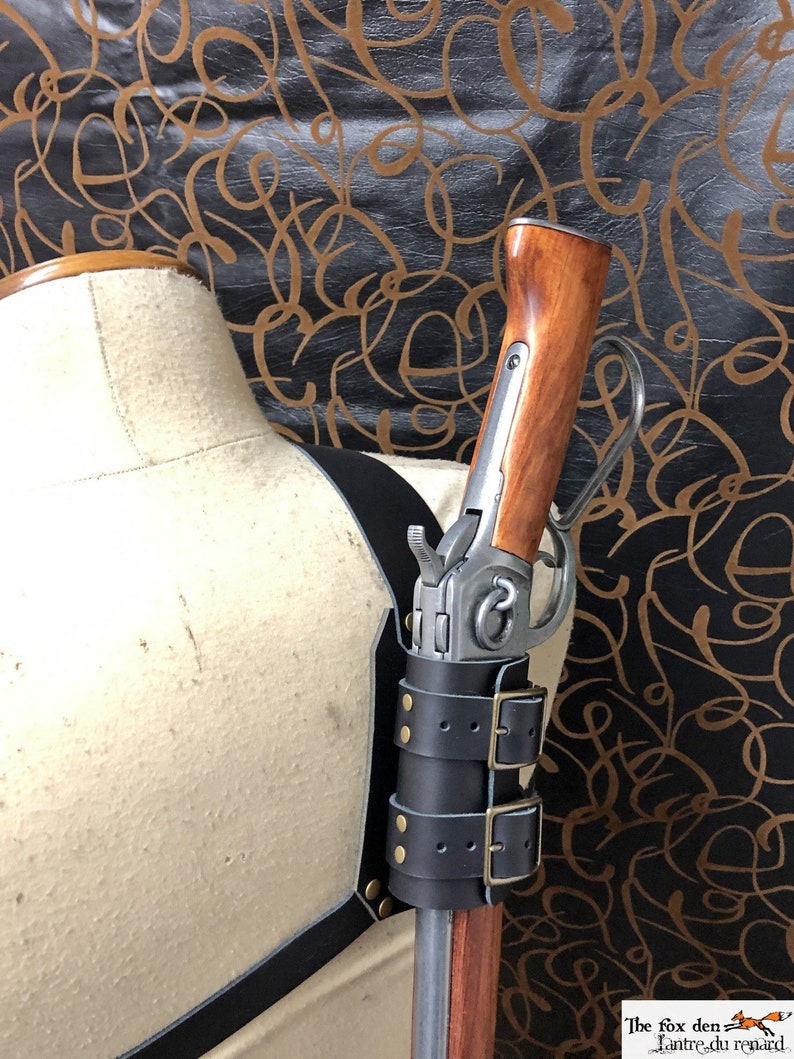 May include: A black leather gun holster with two buckles and brass studs. The holster is attached to a mannequin and holds a long gun with a wooden stock.