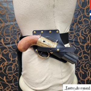 Könnte beinhalten: Ein schwarzes Lederholster mit einer goldenen und braunen Pistole. Das Holster ist an einem schwarzen Gürtel mit silbernen Nieten befestigt.