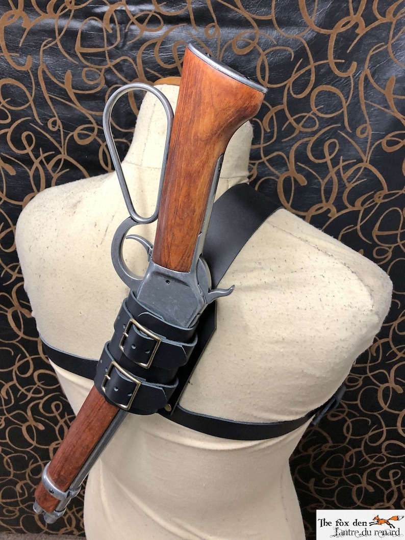 May include: A leather shoulder holster with a rifle attached. The rifle has a wooden stock and a metal barrel. The holster has two buckles and is made of black leather.