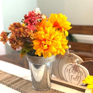 Może przedstawiać: Srebrny metalowy wazon wypełniony żywymi pomarańczowymi i żółtymi sztucznymi kwiatami. Wazon jest ozdobiony sznurkiem wokół góry. W tle dekoracyjne drewniane dynie z napisem "Happy Fall".