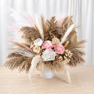May include: A floral arrangement in a white vase featuring dried pampas grass, white and pink feathers, and wooden flowers in shades of white, cream, and soft pink. The bouquet includes various textures of dried grasses and small filler flowers.