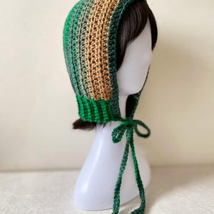 Peut inclure: Un bonnet en crochet présentant des rayures verticales dans des tons vert forêt et beige. La pièce possède un bord côtelé vert le long de la base et comprend deux longs liens fins en crochet vert qui pendent sous le menton.