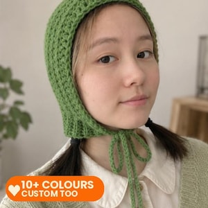 May include: A green crochet bonnet featuring a textured stitch pattern and long chin ties. The bonnet is crafted from yarn and includes an orange graphic overlay with a white heart icon and the text 10+ COLOURS CUSTOM TOO.