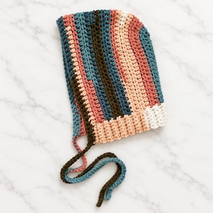 May include: A crochet bonnet featuring vertical stripes in shades of teal, dark olive green, dusty rose, and cream. The headpiece includes a ribbed trim along the front edge and two long, braided ties in dark olive green and teal for fastening.