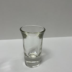May include: A clear, empty shot glass with a thick base and a slightly flared rim. The glass is transparent, allowing light to pass through. It is designed for serving small portions of liquor.