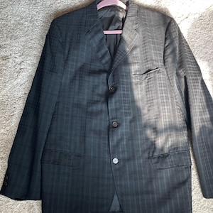 May include: A dark charcoal gray blazer featuring a subtle black plaid pattern, a notched lapel, three front buttons, a single chest pocket, and two lower flap pockets. The garment is fully lined and includes three small buttons on each sleeve cuff.