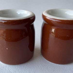 Könnte beinhalten: Zwei kleine, braune Keramiktöpfe mit glänzender Oberfläche. Jeder Topf hat einen weißen Innenraum und einen leicht ausgestellten Rand. Die Töpfe dienen wahrscheinlich zum Servieren einzelner Portionen.