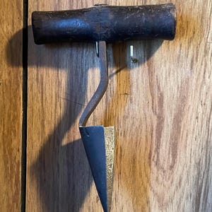 May include: A vintage corkscrew with a dark wooden handle and a curved metal arm. The pointed end is a dark blue color with a gold-colored side. The tool is designed for removing corks from bottles.