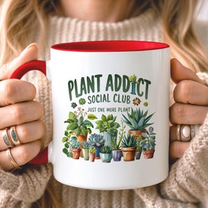 May include: A white ceramic mug with a red handle and interior rim. The front features a colorful illustration of various potted succulents and houseplants with the text "PLANT ADDICT SOCIAL CLUB" and "JUST ONE MORE PLANT" printed in green.