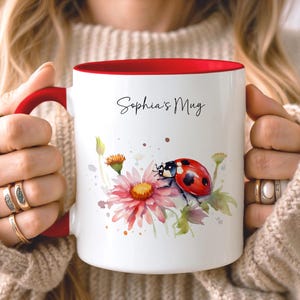 May include: A white ceramic mug with a red interior and handle, featuring a watercolor illustration of a ladybug on a pink flower. The text 'Sophia's Mug' is printed in black script on the upper portion of the mug, centered above the ladybug.
