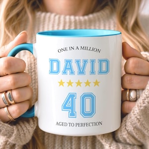 May include: A white ceramic mug with a bright blue handle and interior rim. The front features the text "ONE IN A MILLION", the name "DAVID" in large blue block letters, five yellow stars, the number "40" in blue, and "AGED TO PERFECTION".
