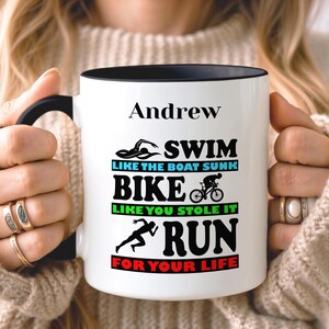 May include: White ceramic mug with a black interior and handle. The mug features the name "Andrew" above the words "SWIM, BIKE, RUN" with related graphics and phrases. The mug is held by a person wearing rings.